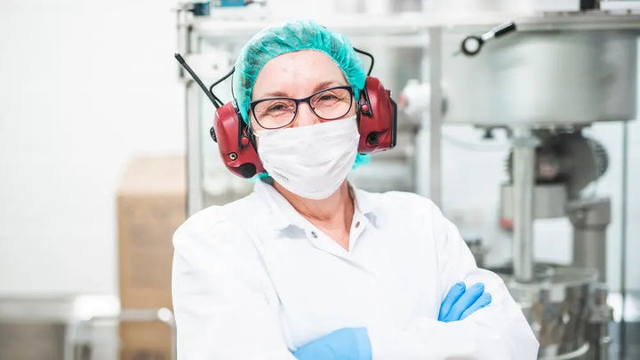 En dame i labfrakk, hårnett, hansker og med hørselsvern på hodet, ser støtt inn i kamera med hendene i kryss. Hun vises fra brystet og opp og står inne i en lab hvor det produseres kosttilskudd