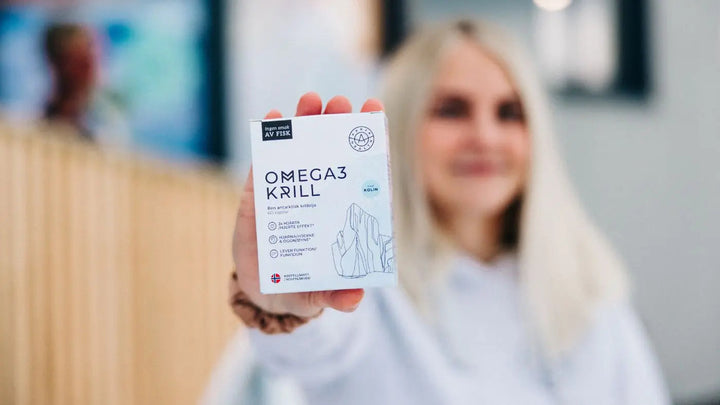 En kvinne med langt, lyst hår i hvit genser holder oppe en boks med kosttilskudd av Omega 3 Krill. Hun er diffus i bakgrunnen og hun er ute