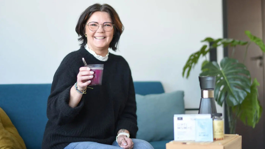 En smilende dam med kort, mørkt hår og en svart genser på, sitter på en sofa og holder et glass med kosttilskudd i. Foran henne står det en boks med kosttilskudd.
