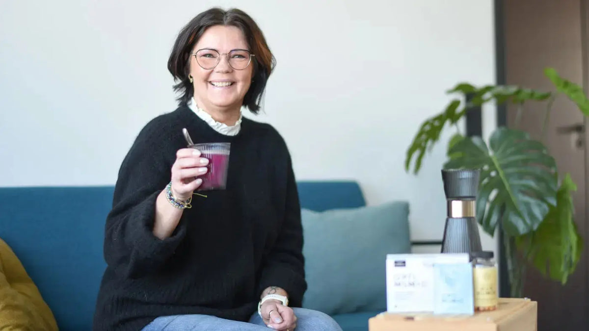 En smilende dam med kort, mørkt hår og en svart genser på, sitter på en sofa og holder et glass med kosttilskudd i. Foran henne står det en boks med kosttilskudd.