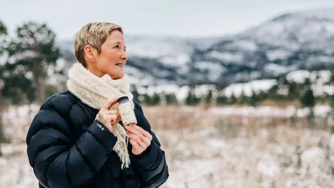 En dame med kort, blondt hår står foran et snødekt område fra hoften og opp. Hun har på en svart boblejakke og et hvitt skjerf, i hånden har hun en boks med kollagen