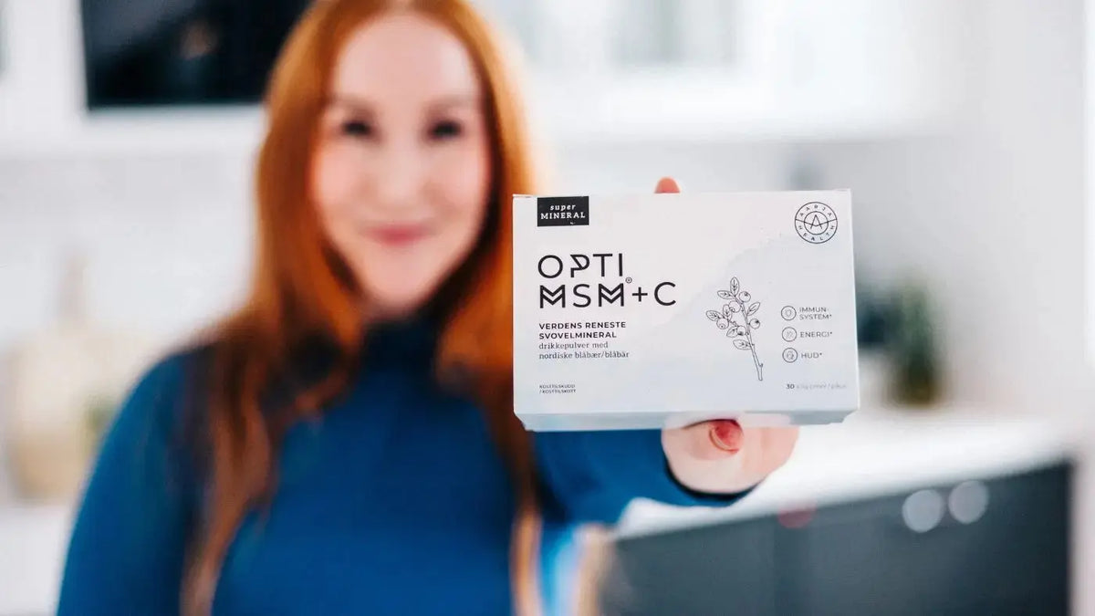 En dame kledt i blått og med langt rødt hår står ufokusert fra brystkassa og opp, mens hun viser fram en boks med OptiMSM + C