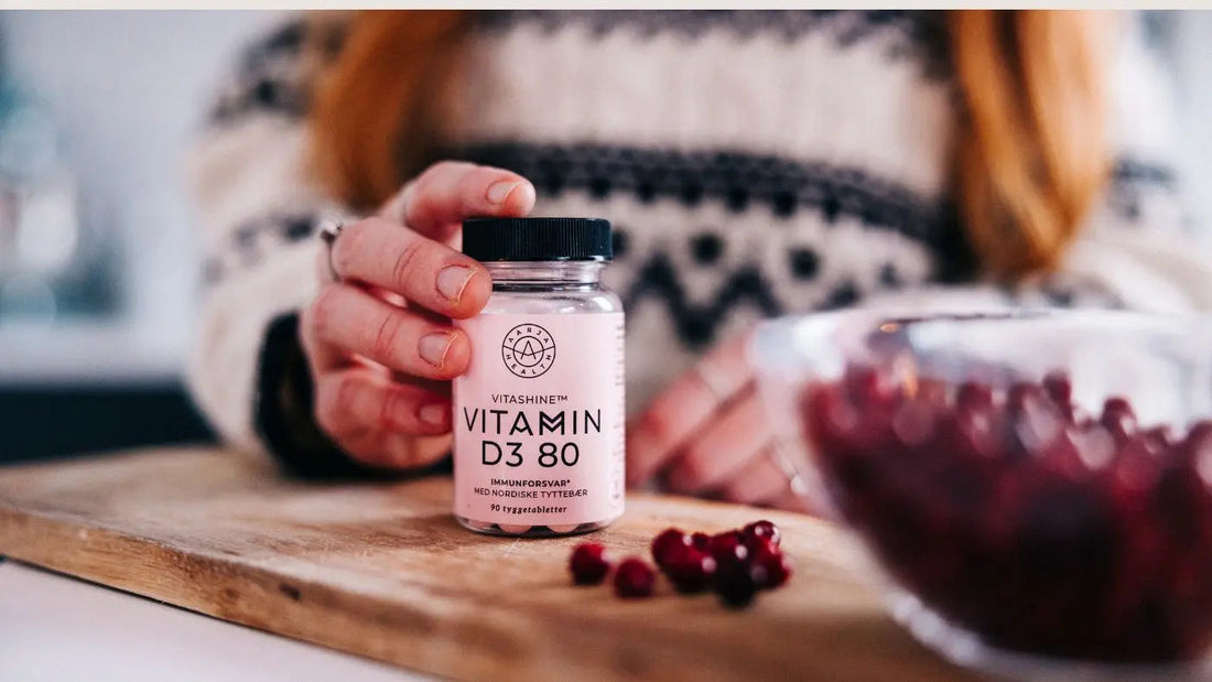 Inne på en kjøkkenbenk holder en kvinne en boks med D-vitamin sammen med tyttebær, ufokusert bakgrunn. står det tre bokser med kosttilskudd, en blå, en rosa og en gul i fokus, bakgrunnen er ufokusert