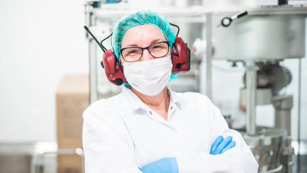 En dame i labfrakk, hårnett, hansker og med hørselsvern på hodet, ser støtt inn i kamera med hendene i kryss. Hun vises fra brystet og opp og står inne i en lab hvor det produseres kosttilskudd